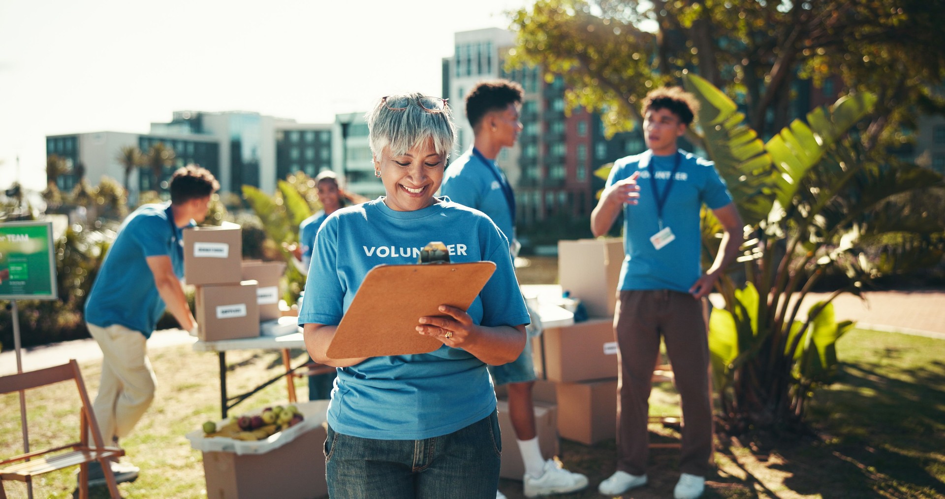 Happy, volunteers and mature woman with clipboard, charity and inventory for donations, smile and ngo. Outdoor, team and coordinator with checklist for info, community service and boxes for nonprofit